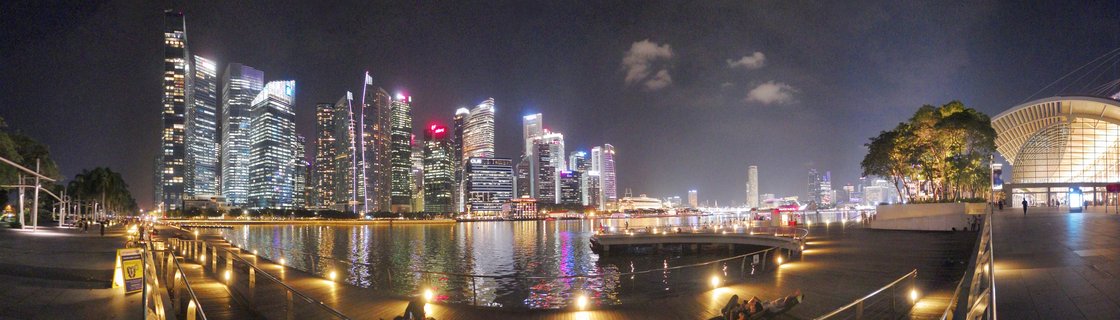 Nighttime panoramic view of Singapore's illuminated CBD skyscrapers across Marina Bay with waterfront promenade and light reflections on water