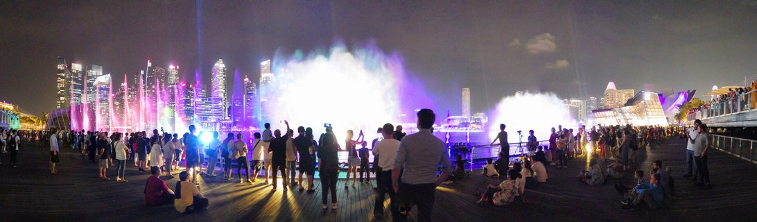 Panoramic night view showing Spectra light and water show at Marina Bay Singapore with blue and purple illuminated fountains, laser beams, crowds of spectators along waterfront railing, and illuminated CBD skyscrapers with reflections on bay water