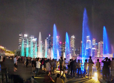 Night view of colored fountain water jets at Marina Bay Singapore with crowd of spectators watching and illuminated skyscrapers in background