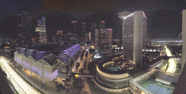 Aerial night photograph of Singapore's Marina Bay showing illuminated skyscrapers, the Pan Pacific Hotel tower, Suntec Convention Centre with purple lighting, and light trails from traffic on streets below