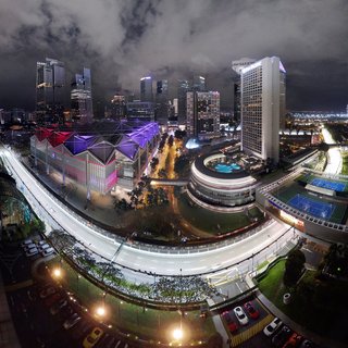 Night aerial photograph of Singapore's Marina Bay showing the Formula 1 street circuit with vehicle light trails, Suntec City Convention Centre with colorful roof lighting, and illuminated skyscrapers against a cloudy night sky