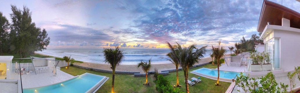 Luxury beachfront villa with two swimming pools, palm trees with uplighting, white modern buildings, sandy beach, ocean waves, and colorful sunset sky in Phuket Thailand