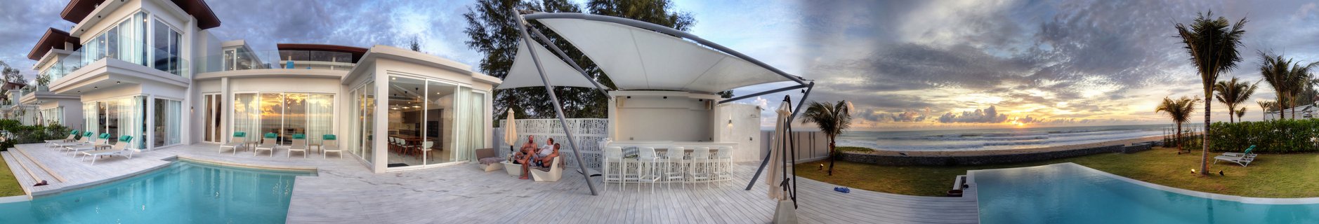 Panoramic view of a white modern beachfront villa with two blue infinity pools, outdoor dining area with people seated, palm trees, and ocean sunset in Phuket Thailand