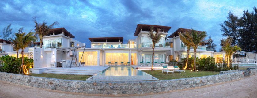 Luxury white villa complex with multiple buildings, large glass windows with warm interior lights, swimming pool in foreground, palm trees, tropical landscaping, dramatic cloudy blue twilight sky, Phuket Thailand