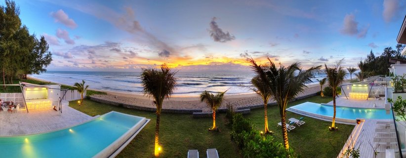 Panoramic view of a luxury beachfront resort in Phuket with two infinity pools, palm trees, white modern buildings with outdoor bars, green lawn, sandy beach, and ocean at sunset with colorful sky