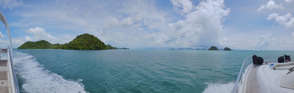 View from a luxury yacht showing turquoise tropical ocean water with white boat wake, limestone karst islands covered in green vegetation in the distance, and dramatic white cumulus clouds in blue sky over Phang Nga Bay, Thailand