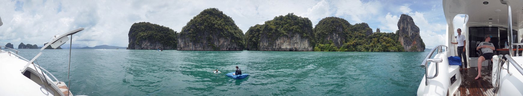 Panoramic view of turquoise bay water with green-covered limestone rock formations, a person kayaking in blue kayak, and white boat with passengers visible on the right side