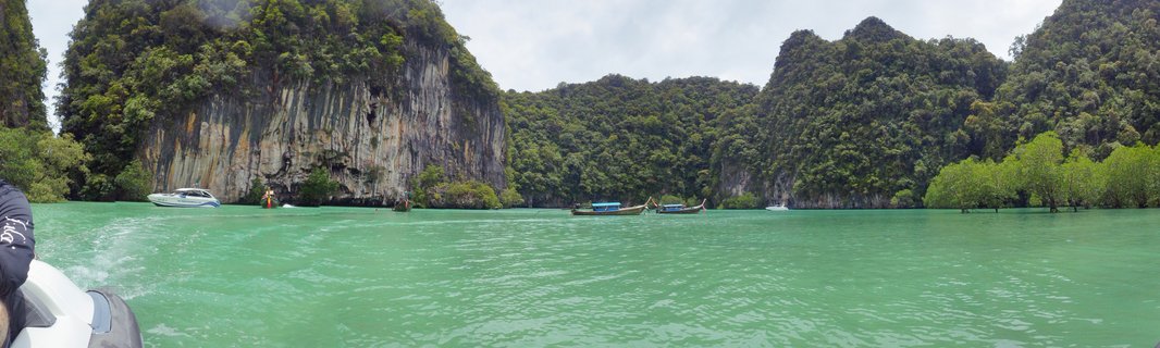 Panoramic view of traditional Thai longtail boats and white speedboats on turquoise water with large limestone cliffs covered in green vegetation in the background