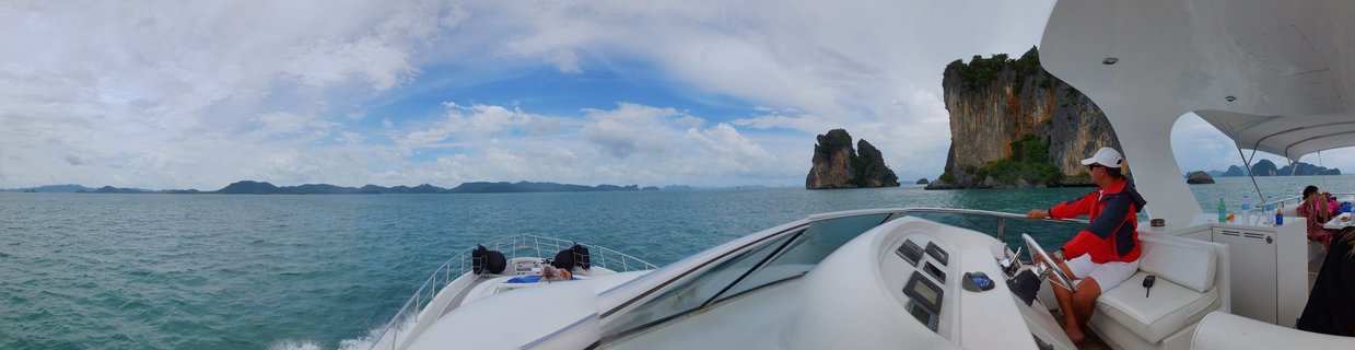 A person in a red jacket and white cap steers a white luxury speedboat through turquoise tropical waters in Phang Nga Bay, Thailand, with dramatic limestone karsts and cliffs rising from the sea under a blue sky with white clouds.
