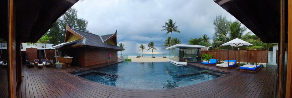 Luxury beachfront villa in Phuket with swimming pool on wooden deck, traditional Thai-style building, palm trees, daybeds with blue cushions, sandy beach and ocean under cloudy sky