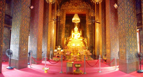 Golden Buddha statue seated on ornate pedestal inside Thai Buddhist temple with decorated columns, red carpet, and flower offerings