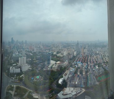 High-angle aerial photograph of Nanjing city showing dense urban development with skyscrapers, apartment buildings, tree-lined streets, and a sports field under cloudy overcast sky