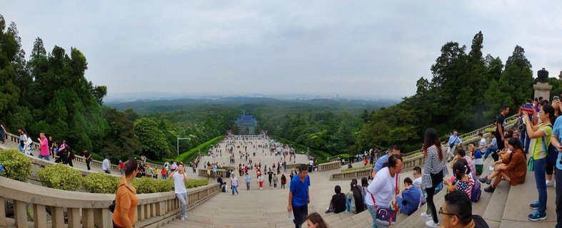 Wide panoramic view of tourists walking on grand stone staircase leading to Sun Yat-sen Mausoleum with blue-tiled roof building visible in distance, surrounded by green forest under overcast sky