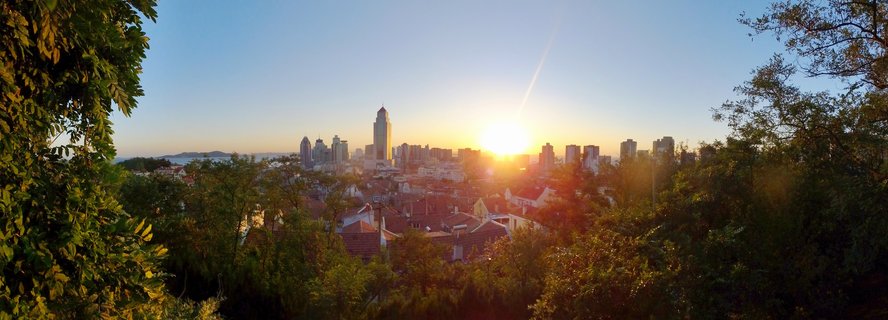 Panoramic view of Qingdao city skyline at sunset with modern skyscrapers, traditional red-roofed buildings, and green trees in foreground