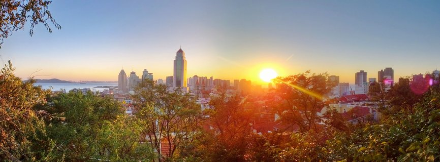 Panoramic view of Qingdao China city skyline at sunset with modern skyscrapers, red-roofed buildings, green trees in foreground, and sun creating golden glow over urban landscape