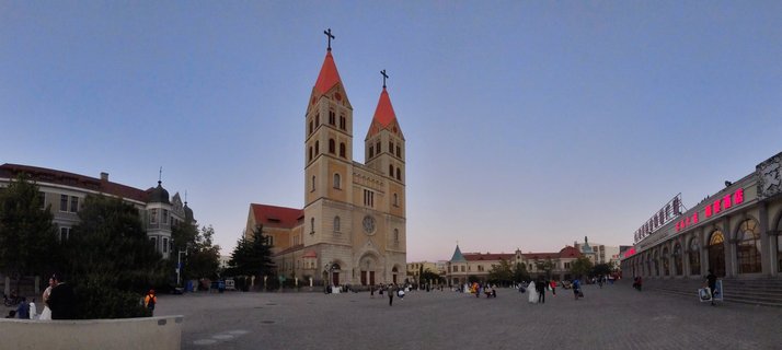 A Catholic cathedral with two tall towers topped by red roofs and crosses, standing in a large paved public square with people walking around in Qingdao, China