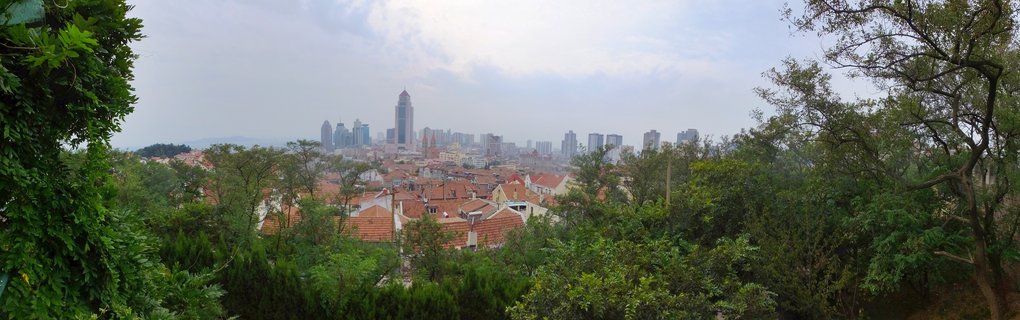 Cityscape of Qingdao China showing red-tiled roof buildings in foreground with modern skyscrapers in distance, framed by green trees on both sides under cloudy sky