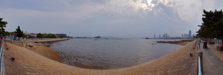 Panoramic waterfront view showing a curved stone seawall, calm bay water with small boats, distant city skyline with modern buildings, tree-lined promenade with pedestrians, and cloudy sky over Qingdao, China