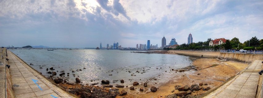 Panoramic coastal view of Qingdao city skyline with modern skyscrapers, rocky beach, concrete seawall, and cloudy sky over Yellow Sea waters
