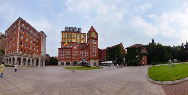 Wide view of Tsingtao Brewery Museum showing red brick German colonial buildings with clock tower, brewing silos, paved plaza with visitors, and green lawn areas under partly cloudy sky