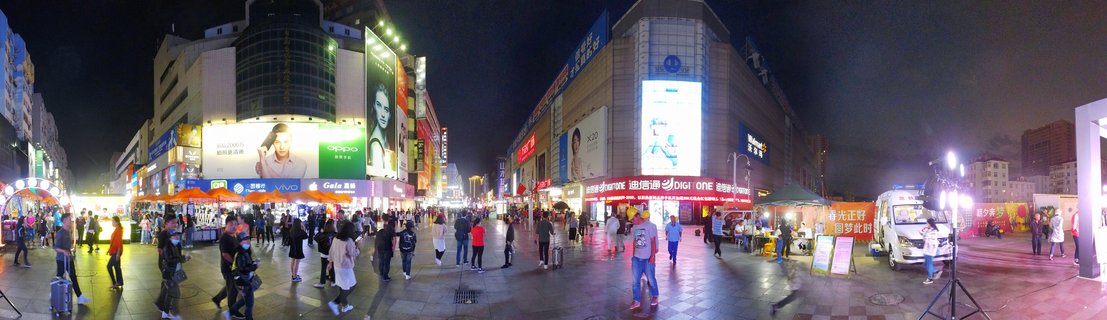 Wide night photograph of a crowded pedestrian shopping street in Qingdao China with bright LED billboards, multi-story buildings, street vendors, and people walking on wet reflective pavement