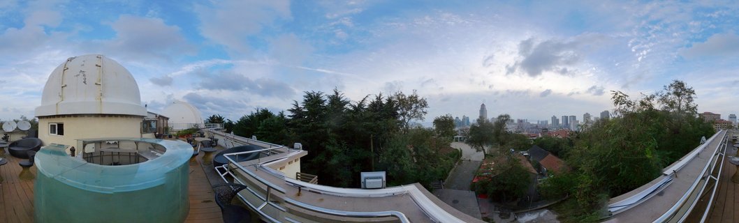 Rooftop view showing white observatory domes on a wooden terrace with curved railings, green trees below, and Qingdao city skyline with skyscrapers in the distance under a blue sky with scattered clouds.