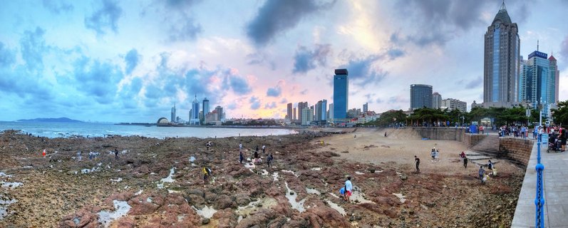 Panoramic coastal cityscape showing Qingdao's skyline with modern skyscrapers, rocky beach with tide pools in the foreground, people exploring the shore, and dramatic cloudy sky at sunset