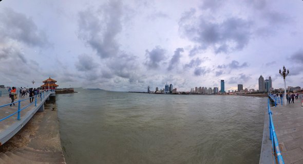 Panoramic view of Zhan Qiao pier with blue railings extending into Qingdao Bay, traditional Chinese pavilion on the left, modern city skyline across the water, people walking on the pier under cloudy sky