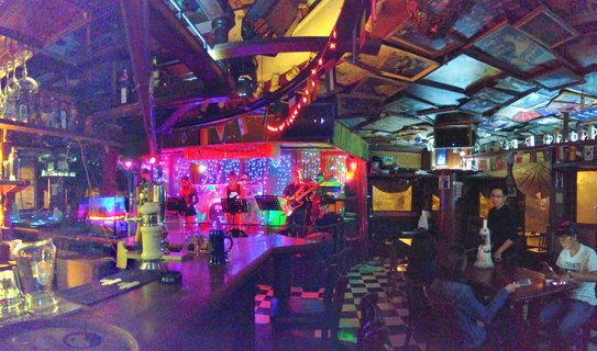 Interior view of a bar with a live band performing on a stage lit by pink, blue and green LED lights. Patrons sit at tables in the foreground while framed pictures decorate the ceiling.