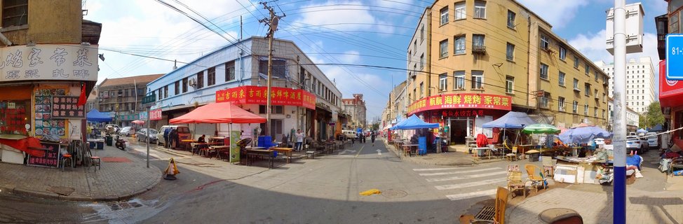 A busy street scene in Qingdao, China showing outdoor food vendors with colorful tents, restaurant signage in Chinese characters, pedestrians walking, and multi-story buildings on both sides of the road under a blue sky