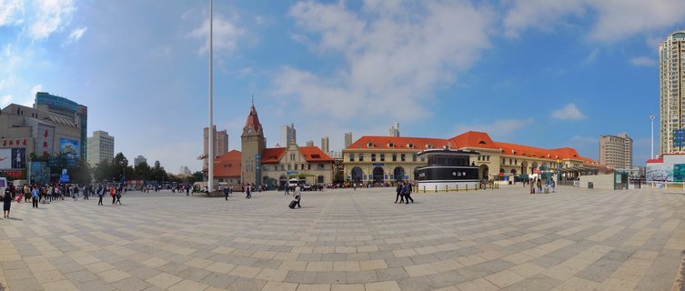 Wide view of Qingdao Railway Station with red-tiled roofs and clock tower, surrounded by modern buildings, with people walking across a large paved plaza under blue sky with clouds