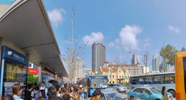 Crowded bus station with people waiting under shelter, blue and white buses, light blue taxis, modern cylindrical skyscraper and European-style buildings in background, clear blue sky