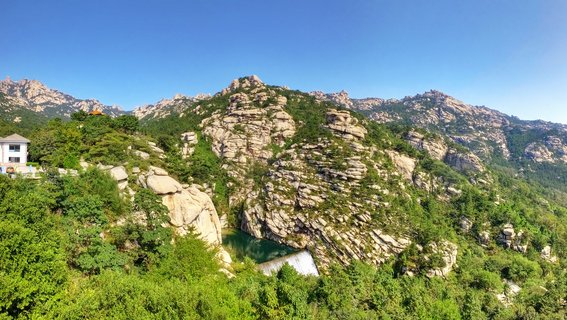 Panoramic view of Laoshan Mountain showing weathered granite rock formations covering hillsides with dense green trees, a white building on the left, and clear blue sky above