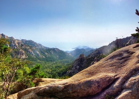 Panoramic mountain landscape showing rocky granite formations and dense green forest on Laoshan Mountain in Qingdao, China, with cable car gondolas visible on the right side and coastal ocean view with islands on the distant horizon under clear blue sky