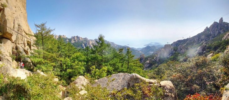 Mountain landscape showing granite rock formations, green pine trees, cable cars suspended on wires, and hikers on a trail with ocean visible in the distance