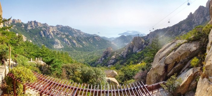 Panoramic view of cable cars suspended over mountain valley with rocky peaks, green forest, and distant ocean view in Qingdao China