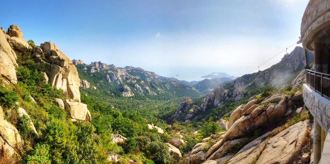 Panoramic mountain landscape showing large granite rock formations, green forest-covered slopes, cable car lines with gondolas, stone observation structure on the right, and ocean with islands in the distant background under clear blue sky