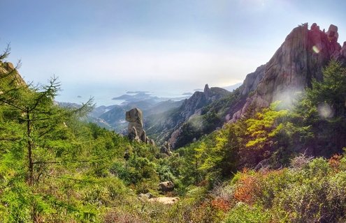 Mountain landscape showing steep granite rock formations and cliffs covered with green coniferous trees, overlooking a coastal bay with islands visible in hazy blue distance under clear sky