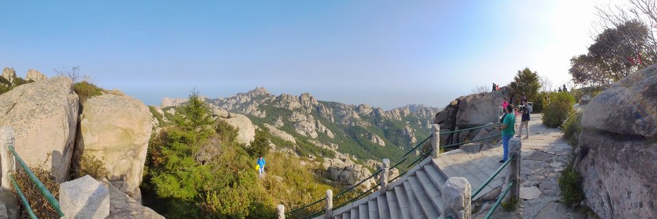 Panoramic view of stone pathway with green railings winding through granite boulders and evergreen trees at Laoshan Mountain, with tourists walking and distant mountain ridges under clear blue sky