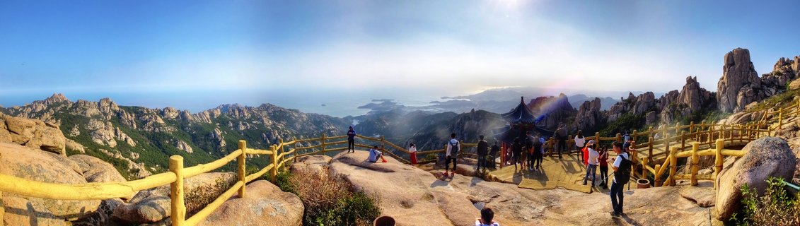Panoramic view from Mount Lao showing tourists at a viewing platform with traditional Chinese pavilion, wooden railings, rocky mountain peaks, and Yellow Sea coastline in the distance