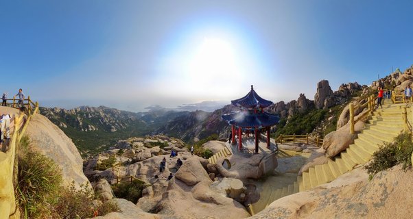 Panoramic view of a traditional Chinese pavilion with blue curved tile roof and red columns on a granite mountain summit. Stone steps and wooden railings lead to the structure with tourists. Rolling mountain peaks with green vegetation extend to distant Yellow Sea horizon under clear blue sky with bright sun.