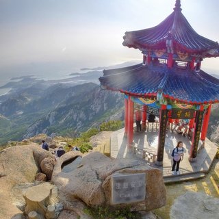 A traditional Chinese pavilion with blue tiled roof and red pillars sits on a rocky mountain peak, with tourists resting inside while overlooking a hazy coastal landscape of mountains and sea