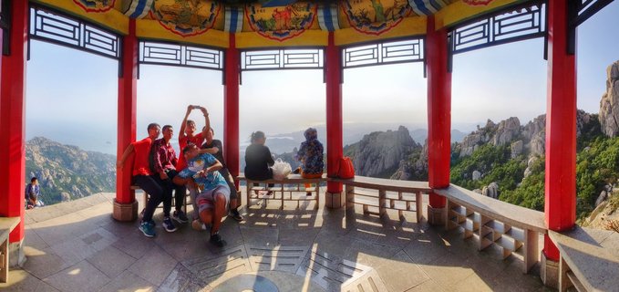 Group of tourists sitting and taking photos inside a traditional Chinese pavilion with red columns, overlooking rocky mountain peaks and distant ocean coastline at Laoshan near Qingdao, China