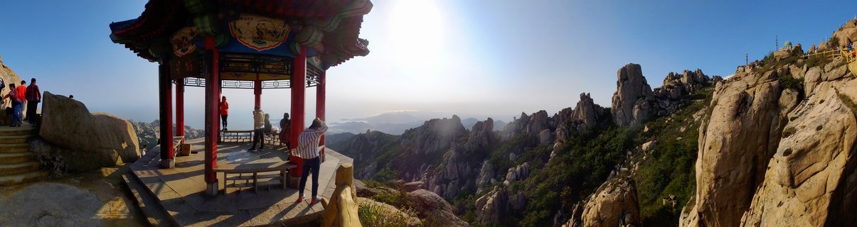 Panoramic view of a traditional Chinese pavilion with red columns on a mountain summit, surrounded by rocky granite peaks and distant hazy mountains under a clear blue sky with tourists visible on the stone platform