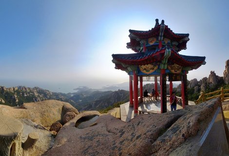 Traditional Chinese pavilion with red columns and ornate multi-tiered roof on mountain peak, with tourists inside, granite boulders in foreground, and coastal mountain landscape in background under clear blue sky