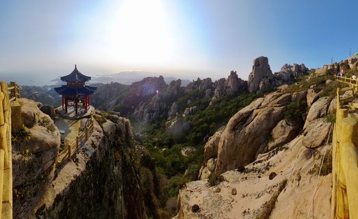 Traditional Chinese pavilion with curved blue roof on rocky mountain peak with wooden railings, granite formations, green vegetation, and distant coastal view under bright sun