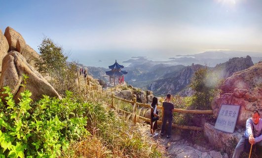 Panoramic mountain landscape showing traditional Chinese pavilion with blue roof on rocky peak, wooden railings along stone pathway, tourists viewing distant coastline and islands under bright sunlight near Qingdao China