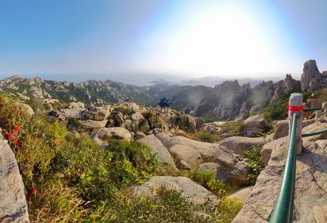 Panoramic view of Laoshan Mountain in Qingdao showing rocky granite peaks, green vegetation, a traditional Chinese pavilion with blue roof, hiking trails with tourists, and the Yellow Sea coastline in the distance under bright afternoon sun