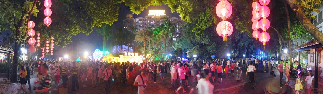 Night scene of a crowded public square in Guangzhou with red Chinese lanterns hanging from trees, people walking and celebrating during Mid-Autumn Festival