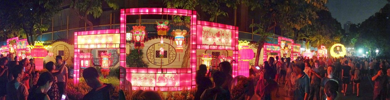 Night scene showing crowds of people walking among large illuminated pink traditional Chinese lantern structures with ornate hanging lanterns at an outdoor festival